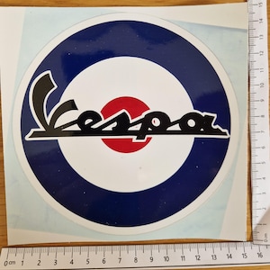 May include: A circular sticker with the Vespa logo in black, set against a white and red bullseye design, encircled by a blue ring. The sticker is on a white backing and is about 14 cm in diameter.