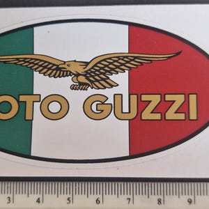 May include: Oval sticker with the Moto Guzzi logo, featuring the Italian flag colours: green, white, and red. A gold eagle is above the gold text "MOTO GUZZI". The sticker is approximately 10 cm long.