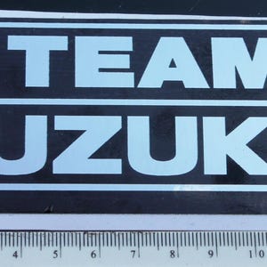 May include: A rectangular sticker with the words "TEAM SUZUKI" in light blue, outlined in white, on a black background. The Suzuki logo, a red "S" inside a red square, is in the upper left corner. A ruler is on the right side.