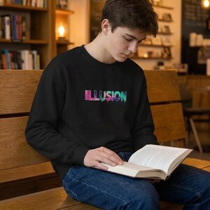 May include: Black crew neck sweatshirt featuring the word "ILLUSION" in a vibrant, dripping font. A person is wearing the sweatshirt and reading a book on a wooden bench. The background shows bookshelves and warm lighting.