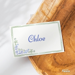 Pode incluir: Um cartão de lugar retangular branco com uma borda verde desenhada à mão e um grupo de flores azuis no canto inferior esquerdo. O nome Chloe está impresso no centro em uma fonte azul. O texto Cosmic Latte Paperie é visível.