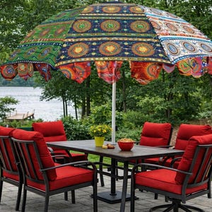 Op de afbeelding: Een kleurrijke terras set met een grote, patroon parasol. De parasol heeft een levendig ontwerp met rode, blauwe, groene en oranje tinten. De set bevat een donkere tafel en rood gevoerde stoelen, waardoor een uitnodigende buitenruimte ontstaat.