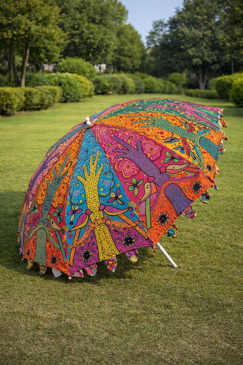 May include: A colorful, open umbrella with a vibrant, patterned design. The umbrella features a white pole and is decorated with hand-like motifs, birds, and floral patterns in shades of blue, orange, pink, and yellow. The umbrella is set on a green lawn.