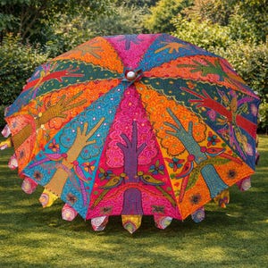 May include: A colorful, open umbrella with a vibrant design. The umbrella features a repeating pattern of hands, trees, and floral elements in shades of pink, orange, blue, and green. The umbrella is set on a grassy lawn.