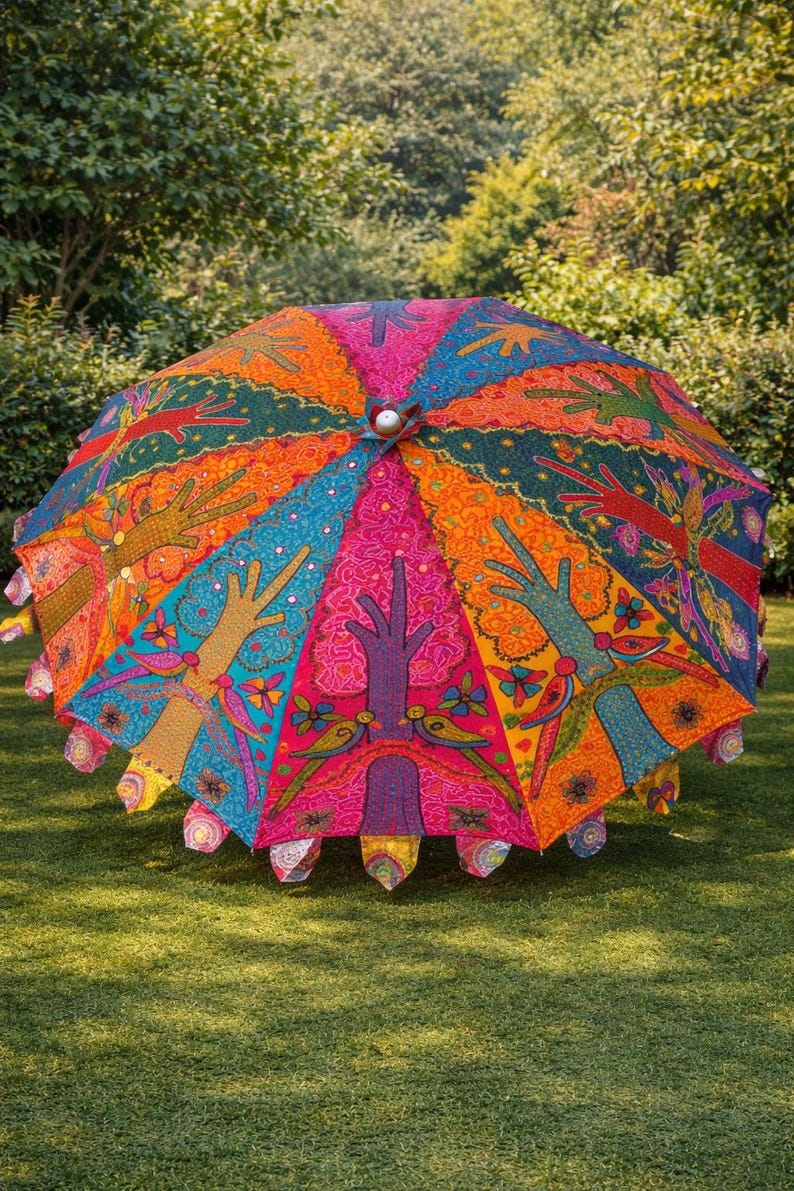 May include: A colorful, open umbrella with a vibrant design. The umbrella features a repeating pattern of hands, trees, and floral elements in shades of pink, orange, blue, and green. The umbrella is set on a grassy lawn.