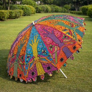 May include: A colorful, open umbrella with a vibrant, patterned design. The umbrella features a white pole and is decorated with hand-like shapes, birds, and floral motifs in shades of orange, pink, blue, and green. The umbrella is set on a green lawn.