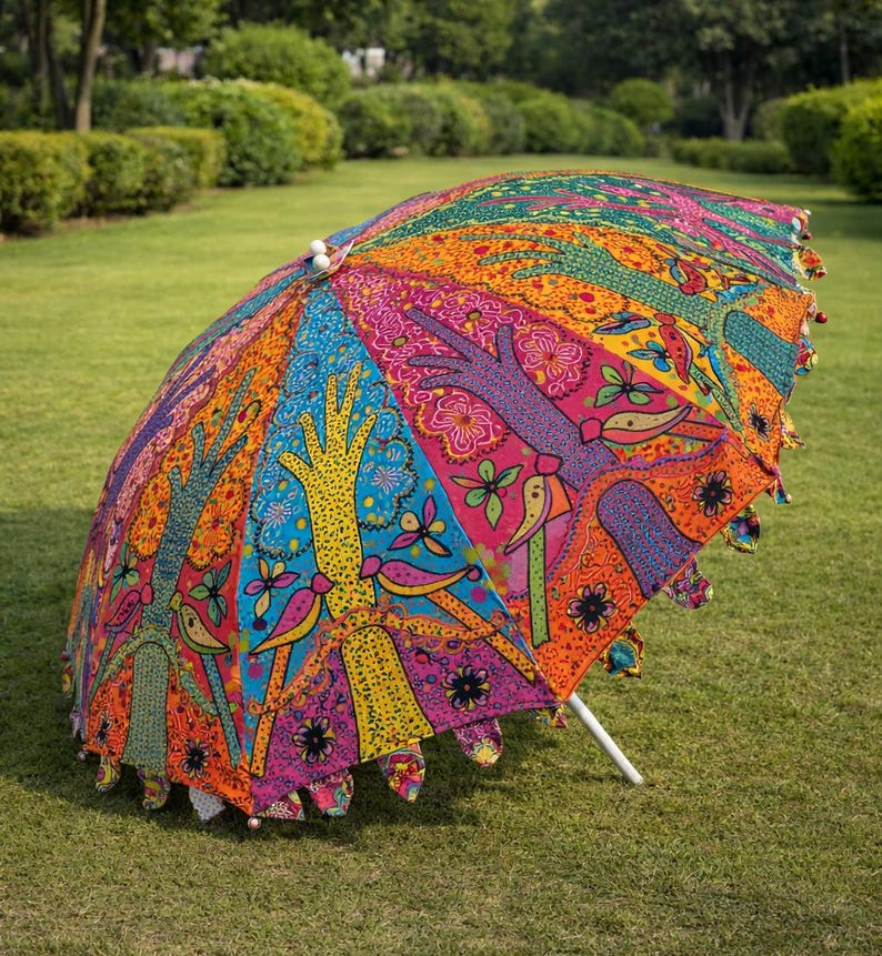 May include: A colorful, open umbrella with a vibrant, patterned design. The umbrella features a white pole and is decorated with hand-like shapes, birds, and floral motifs in shades of orange, pink, blue, and green. The umbrella is set on a green lawn.