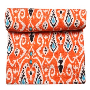 May include: Rolled orange quilt with a repeating pattern of white, black, and turquoise shapes. The quilt is made of a soft, textured fabric and is likely handmade. The design is symmetrical and eye-catching, suitable for a bed or sofa.
