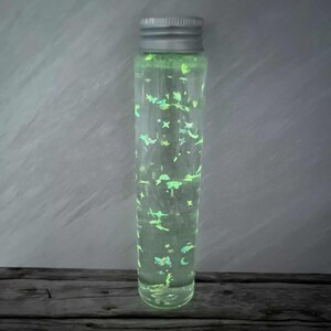 Pode incluir: Um frasco de vidro transparente com tampa prateada, preenchido com líquido verde brilhante e glitter. O glitter inclui estrelas, luas e outras formas. O frasco está sobre uma superfície de madeira escura e um fundo cinzento.