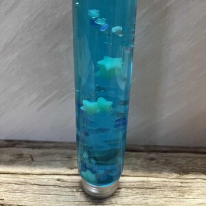 Pode incluir: Uma garrafa sensorial cilíndrica transparente cheia de líquido azul e elementos decorativos. A garrafa contém pequenas formas azuis e transparentes e vários objetos em forma de estrela azul clara. A garrafa tem uma tampa prateada.