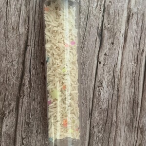 Pode incluir: Um tubo de plástico transparente preenchido com arroz branco e confetes coloridos em forma de estrela. O tubo tem uma tampa de metal prateado. O fundo é uma superfície de madeira com grão vertical.