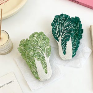 Puede incluir: Dos esculturas decorativas de repollo. Las esculturas son verdes con venas y tallos blancos. Las esculturas de repollo están sobre papel blanco. El fondo incluye una vela, una bandeja y un libro.