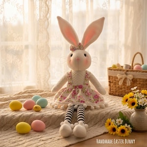 May include: A soft toy Easter bunny with long ears, dressed in a floral dress and a bow. The bunny sits on a blanket with colourful Easter eggs. A basket with eggs and a vase of sunflowers are in the background. Text reads "HANDMADE EASTER BUNNY."