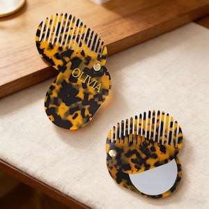 May include: Three tortoiseshell-patterned compact combs with mirrors. One compact features the name "OLIVIA" in gold lettering. The oval-shaped combs have a hinged design, and the comb teeth are white.