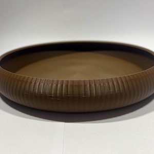 May include: A brown, circular bowl with a textured, ribbed outer edge. The bowl has a wide, shallow design and a smooth interior. The colour is a warm, earthy brown, and the bowl sits on a white surface. This decorative bowl is suitable for home decor.