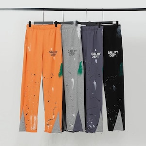 Puede incluir: Cuatro pares de pantalones deportivos en naranja, gris, azul marino y negro, cada uno con detalles de salpicaduras de pintura y el texto "GALLERY DEPT.". Los pantalones tienen un diseño de abertura y cuelgan de una barra negra.