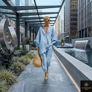 May include: A light blue linen outfit, including a long-sleeved top with a V-neck and matching cropped pants. The outfit is paired with a woven straw tote bag and gold-colored sandals. The background features a modern urban setting with a water feature.