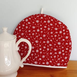 Peut inclure: Un chauffe-thé rouge avec un motif floral blanc et une bordure blanche. Une théière en céramique blanche est partiellement visible à gauche. Le chauffe-thé a une boucle en haut pour l'accrocher.