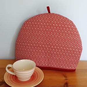 Può includere: Una teiera a fantasia rossa e beige, con rifiniture rosse e un anello. Un set di tazza e piattino coordinati con strisce beige e arancioni sono in primo piano. La teiera è progettata per mantenere il tè caldo.