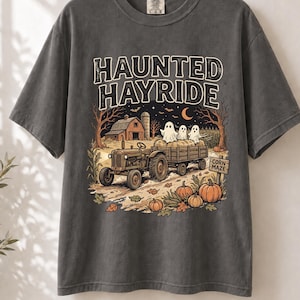 May include: A charcoal gray short-sleeve t-shirt featuring a graphic print with the text "HAUNTED HAYRIDE". The illustration depicts a tractor pulling a wagon filled with three ghosts, a barn, pumpkins, and a sign that reads "CORN MAZE".