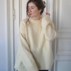 May include: A cream-colored, oversized sweater with a crew neck and long sleeves. The knitwear appears soft and fluffy. The person is wearing a white undershirt. The background is a white door.