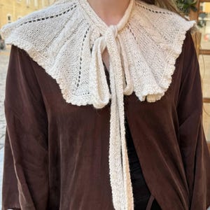 May include: A cream-colored, hand-knitted detachable collar featuring a wide, ruffled design with intricate cable-knit patterns and delicate eyelet details. The collar is secured at the front with long, matching knitted ties that hang down.