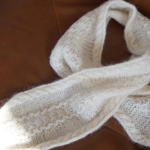 May include: A cream-coloured, knitted scarf with a delicate, openwork pattern. The scarf is made of a soft, fluffy yarn and is draped on a brown leather surface. The scarf's texture and design suggest it is handmade.