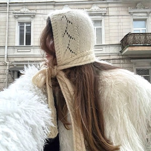 May include: A cream-colored knitted bonnet with a decorative leaf pattern and long ties. The bonnet is made of a soft, textured yarn. The person is wearing a white fluffy coat.