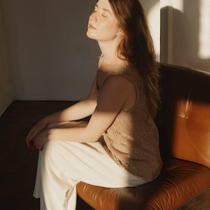 Puede incluir: Una persona con un top de punto beige y pantalones blancos sentada en una silla de cuero marrón. El top sin mangas tiene un patrón texturizado. La silla es de color marrón cálido y la habitación está iluminada por la luz del sol.