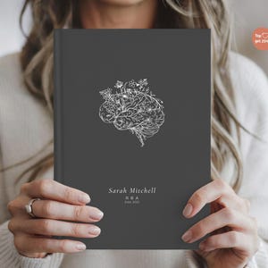 May include: A dark gray journal with a white floral brain design. The journal has the name "Sarah Mitchell" and "RBA Est 2022" printed at the bottom. The journal is held by a person.
