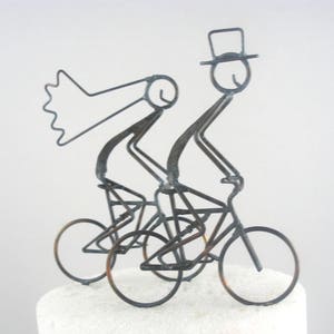 May include: A metal sculpture of a bride and groom riding a tandem bicycle. The bride is wearing a veil and the groom is wearing a top hat. The sculpture is made of thin wire and is painted in a silver color.