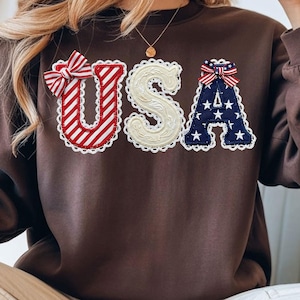 Könnte beinhalten: Ein braunes Sweatshirt mit den Buchstaben "USA" auf der Vorderseite. Das "U" ist rot-weiß gestreift mit einer Schleife, das "S" ist cremefarben und das "A" ist marineblau mit weißen Sternen und einer Schleife. Die Buchstaben sind mit weißer Spitze verziert.