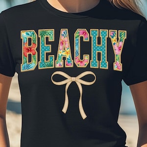 Könnte beinhalten: Schwarzes T-Shirt mit dem Wort "BEACHY" in einem farbenfrohen Blumen- und Polka-Dot-Design. Unter dem Text befindet sich eine beige Schleife. Ein lässiges Kleidungsstück für den Sommer.