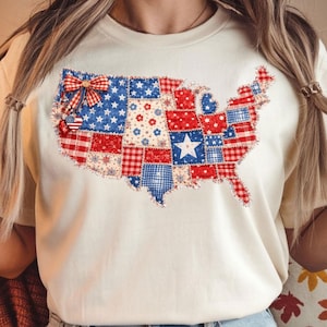 May include: A cream-coloured t-shirt with a patchwork design of the United States map. The map is made up of red, white, and blue fabric squares, with a bow and heart-shaped charm. The design includes stars, floral patterns, and gingham.