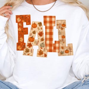 May include: A white long-sleeve sweatshirt featuring the word FALL in large block letters. The letters are filled with patterns of orange autumn leaves, pumpkins, and orange and tan plaid, with a stitched outline around each letter.