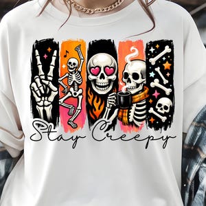 May include: A white t-shirt featuring a graphic print of skeletons, flames, and stars with the text Stay Creepy. The design includes a skeleton hand making a peace sign, a dancing skeleton, a skull with heart eyes, and a skeleton drinking coffee.
