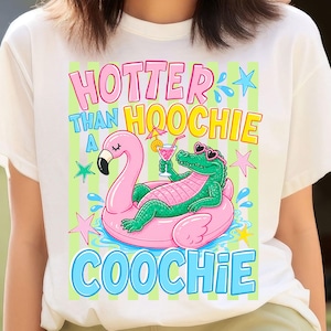 May include: White t-shirt with a colorful graphic. The graphic features a pink flamingo float with a green alligator wearing sunglasses and holding a cocktail. Text reads "Hotter Than Hoochie Coochie."