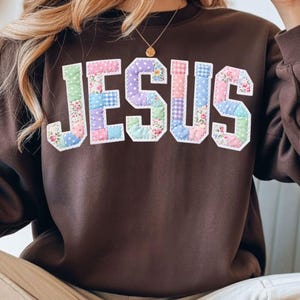 May include: A brown sweatshirt with the word "JESUS" in large, patchwork-style letters. The letters are made of various pastel-colored fabrics, including floral prints, polka dots, and gingham patterns. The sweatshirt has a crew neck and long sleeves.