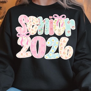 May include: Black sweatshirt with the words "Senior 2026" in a colorful, floral design. The text features pink bows and a variety of patterns, including stripes, flowers, and daisies. A stylish garment for the graduating class of 2026.