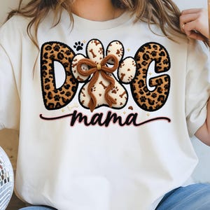 May include: Cream-colored t-shirt with a "DOG MAMA" design. The word "DOG" features leopard print letters. A paw print and a paw-shaped design with a brown bow and bone accents are in the center. The word "mama" is in cursive.