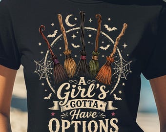 May include: A black t-shirt featuring a graphic of five different broomsticks arranged in a semi-circle above a crescent moon, stars, and spiderwebs. The text reads A Girl's GOTTA Have OPTIONS in a decorative font with small bats surrounding the design.