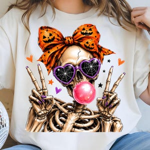 May include: A cream-colored t-shirt featuring a graphic of a skeleton wearing a large orange bow with jack-o'-lantern faces, purple heart-shaped sunglasses with spiderweb patterns, and blowing a bright pink bubble with both hands making peace signs.