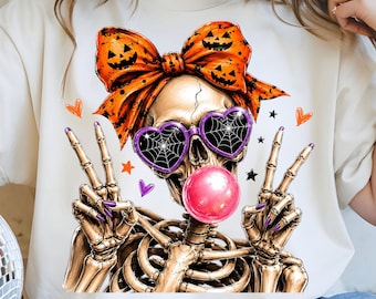 May include: A cream-coloured t-shirt featuring a graphic of a skeleton wearing a large orange bow with jack-o'-lantern faces, purple heart-shaped sunglasses with spiderweb patterns, and blowing a bright pink bubble with both hands making peace signs.