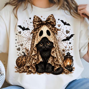 May include: A cream-colored t-shirt featuring a graphic of a black cat wearing a leopard print ghost costume with a matching bow. The design includes a spider web background, leopard print pumpkins, flying bats, and a glowing lantern illustration.