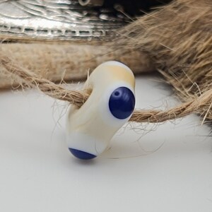 May include: A cream and blue glass bead on a natural fibre cord. The bead has a rounded shape with a dark blue circle on one side and a white base. The background includes a silver metal piece and brown fur. The bead is likely for jewellery making.