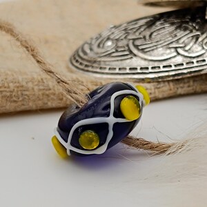 May include: A dark blue glass bead with white and yellow detailing. The bead is oval-shaped, featuring a white grid design and yellow circular elements. A silver-coloured metal clasp and fabric are in the background.