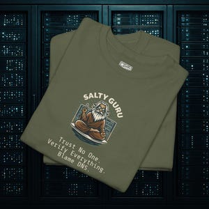 May include: Two olive green t-shirts are stacked. The top shirt features a graphic of a guru with the text "Salty Guru" and the phrases "Trust No One. Verify Everything. Blame DNS." A small white label is visible on the top right of the shirt.