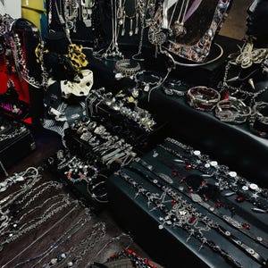 Puede incluir: Una exhibición de varias joyas plateadas, que incluyen collares, pulseras y anillos. Algunas piezas presentan piedras preciosas y elementos decorativos. Las joyas están dispuestas sobre superficies de exhibición negras, mostrando una variedad de estilos y diseños.