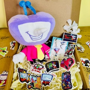 May include: A plush toy shaped like a purple television set with a smiling face, wearing a pink jacket and yellow tie, sits in a cardboard box filled with stickers, a keychain, and cards. The stickers feature cartoon TV characters.