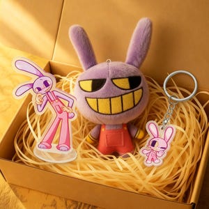 Puede incluir: Una caja de regalo que contiene un conejito de peluche morado con una sonrisa amarilla y un peto rojo, un llavero con un conejito rosa y un expositor acrílico de conejito rosa. La caja está llena de paja.